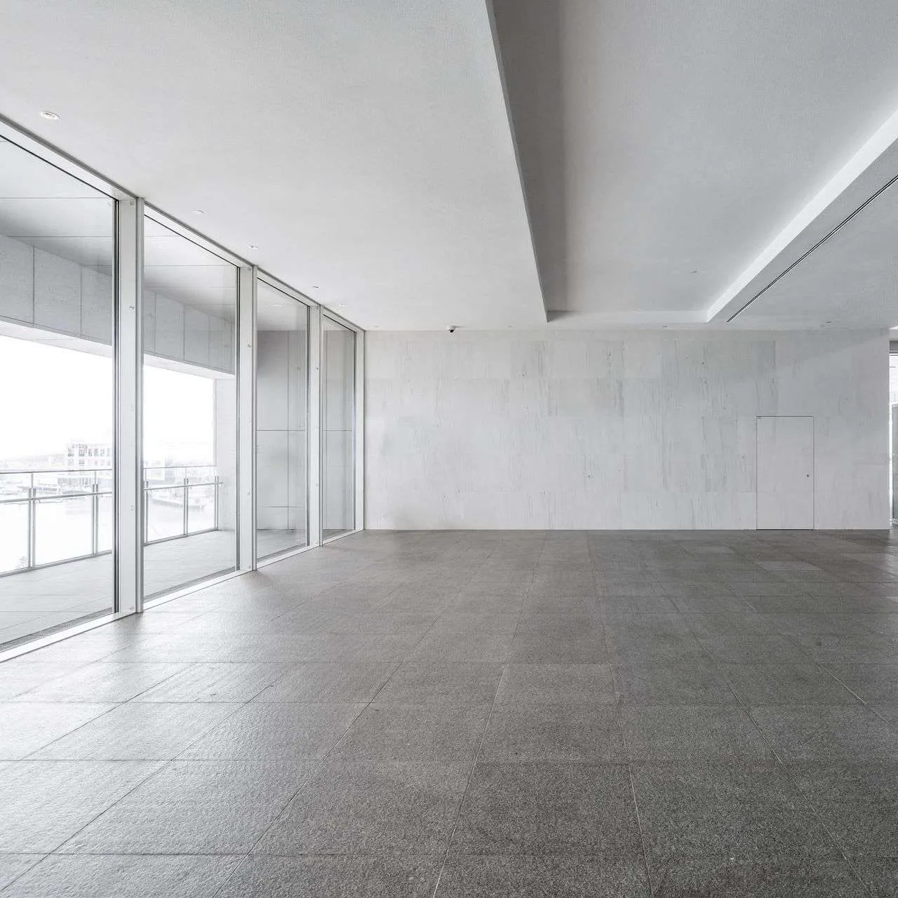 Puste, jasne pomieszczenie biurowe z szarymi płytkami podłogowymi, dużymi oknami z widokiem na miasto i gładkimi, białymi ścianami.