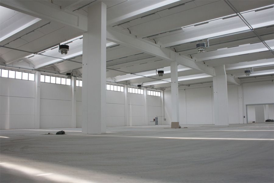 Wnętrze hali magazynowej z białymi ścianami, betonową podłogą, białymi słupami i belkami stropowymi oraz rzędem okien pod sufitem, oświetlone lampami przemysłowymi.