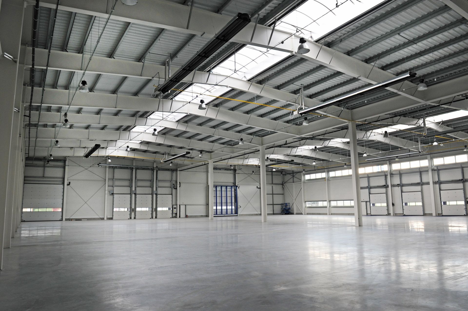 Pusta, jasna hala przemysłowa z betonową posadzką, stalową konstrukcją dachu i szeregiem bram segmentowych.
