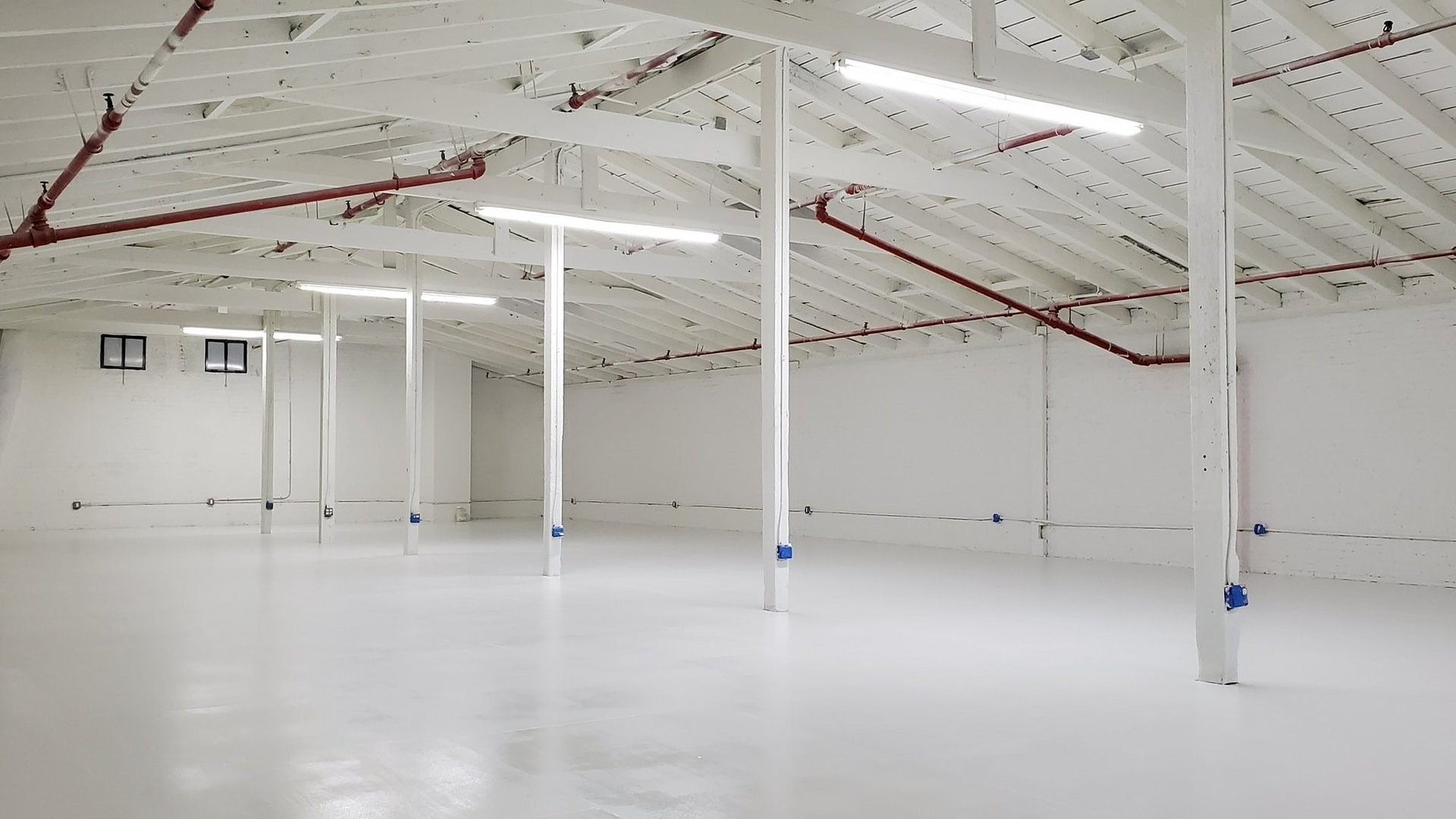 Pomalowane na biało, puste wnętrze magazynu lub biura z drewnianą konstrukcją stropu, czerwonymi rurami i świeżo pomalowaną podłogą z połyskiem.