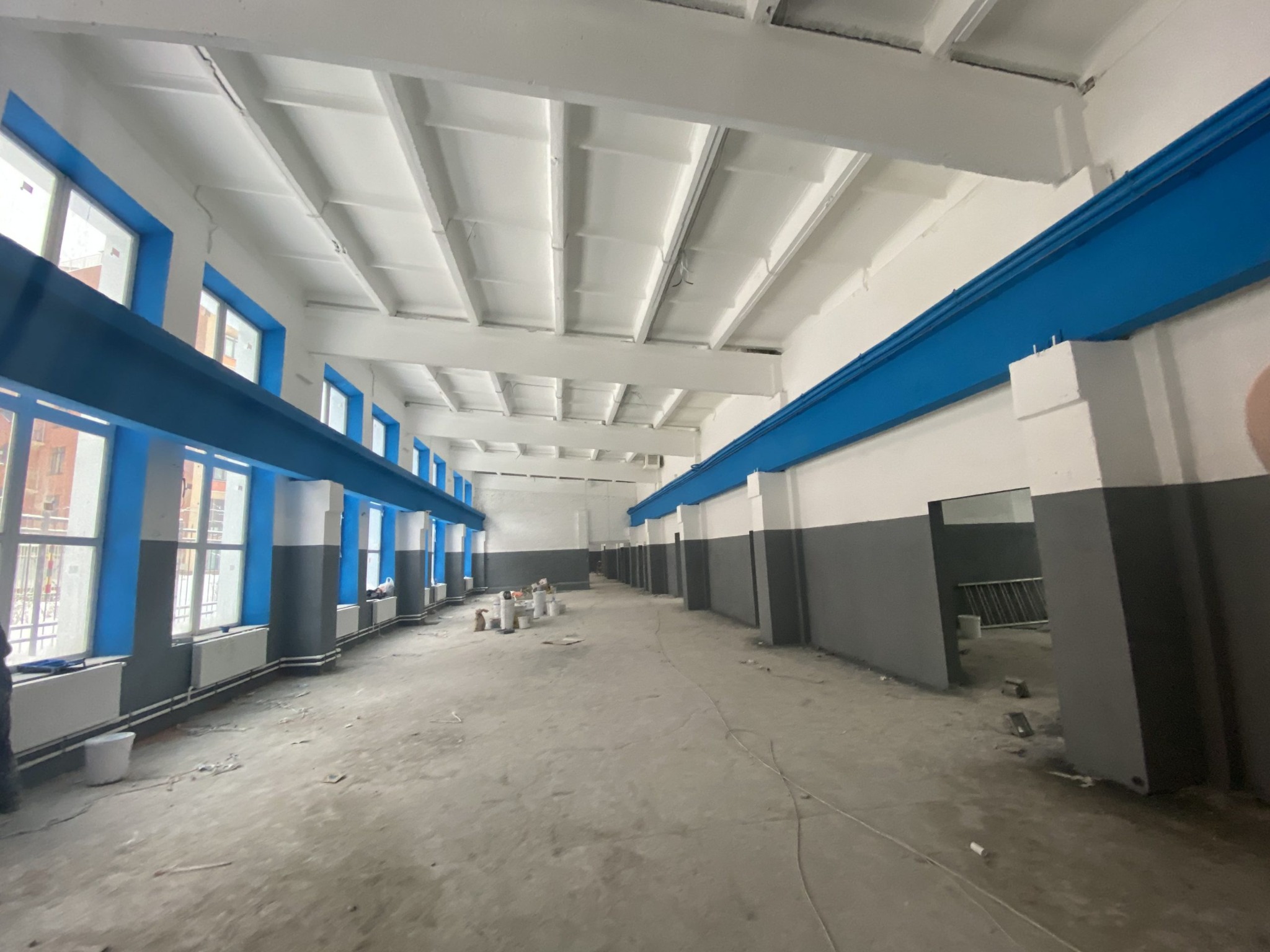 Wnętrze dużego pomieszczenia w trakcie prac malarskich; ściany częściowo pomalowane na szaro, okna i belki na niebiesko, sufit biały; widoczne wiadra z farbą i drabina w tle.