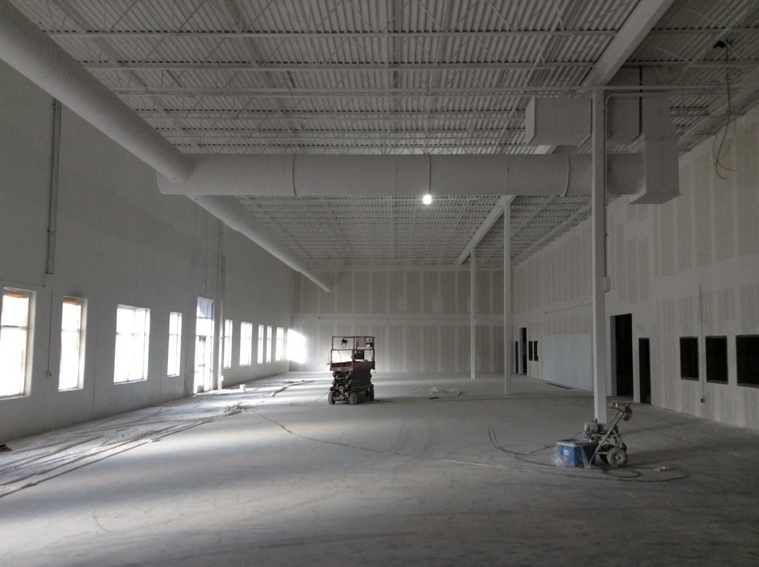 Przestronne wnętrze biurowe w trakcie prac wykończeniowych: surowe ściany z płyt gipsowo-kartonowych, widoczne instalacje wentylacyjne pod sufitem, maszyna do szlifowania posadzki i podnośnik...