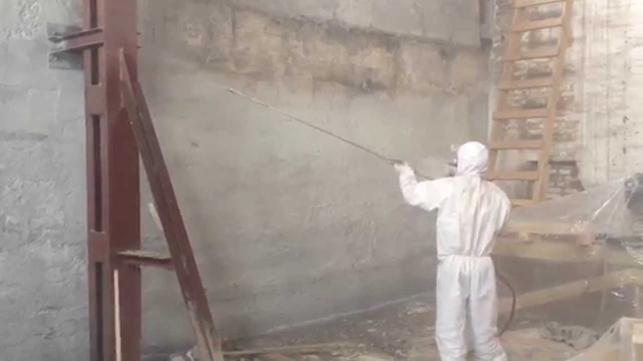 Pracownik w kombinezonie ochronnym natryskuje farbę na surową ścianę wewnątrz budynku, widoczne rusztowanie i drabina.