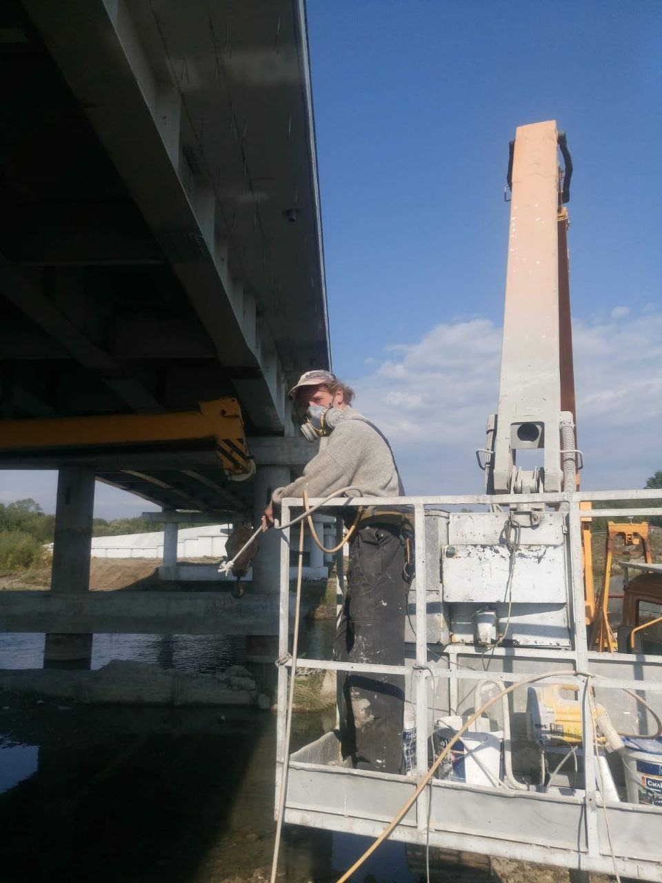 Pracownik w kombinezonie i masce ochronnej maluje konstrukcję mostu z podnośnika koszowego, widoczne farby i narzędzia, w tle rzeka i niebo.