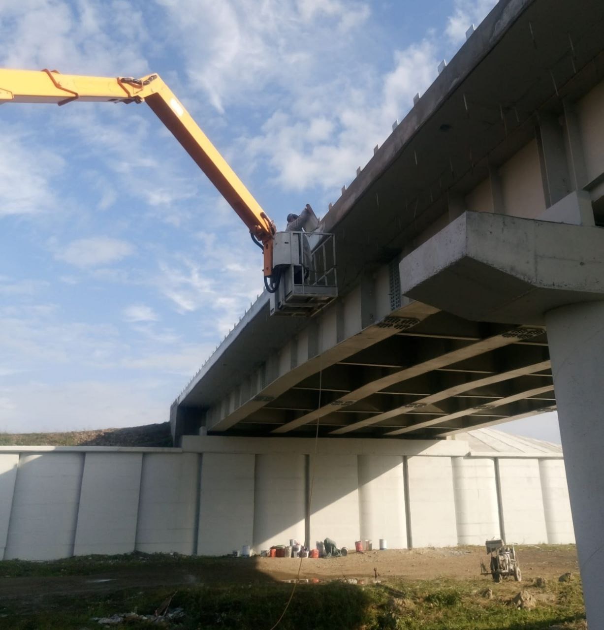Pracownik w podnośniku koszowym aplikuje powłokę ochronną na betonową konstrukcję wiaduktu; widoczne puszki z farbą i agregat malarski.