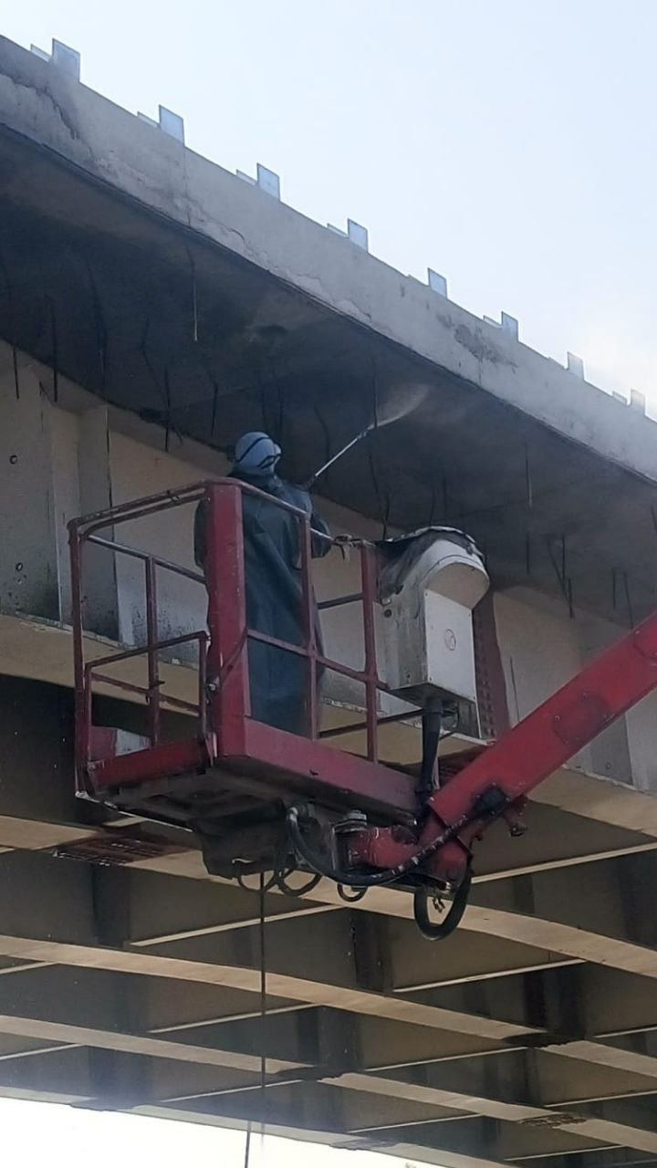 Pracownik w kombinezonie ochronnym i masce, stojący na podnośniku koszowym, czyści konstrukcję betonową mostu za pomocą myjki ciśnieniowej.