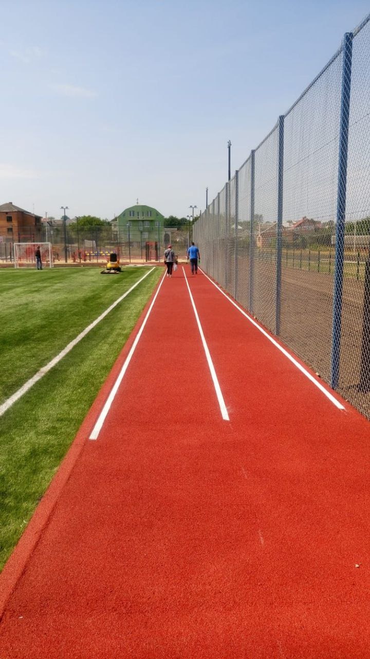 Czerwona bieżnia z białymi liniami na stadionie, z dwoma osobami w oddali i zielonym boiskiem po lewej stronie, oddzielonym białymi liniami, oraz siatką ogrodzeniową po prawej stronie.