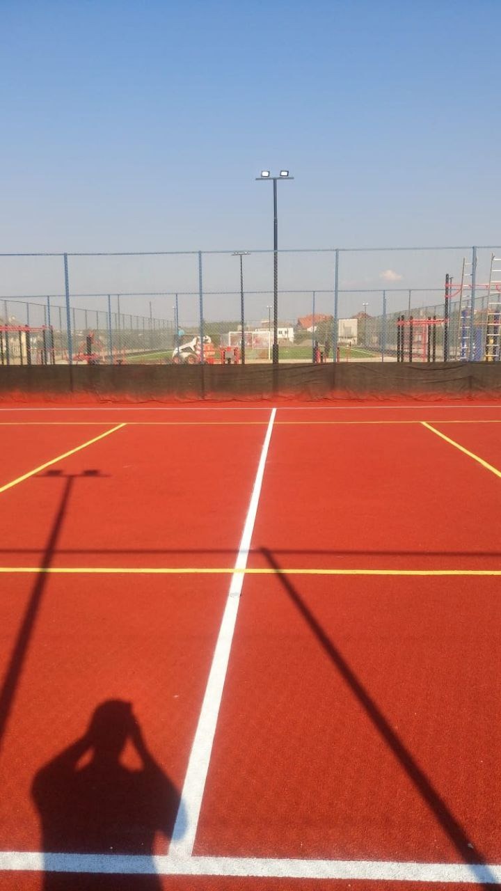 Czerwona nawierzchnia boiska sportowego z białymi i żółtymi liniami, cień fotografa w rogu kadru, siatka ogrodzeniowa w tle, błękitne niebo