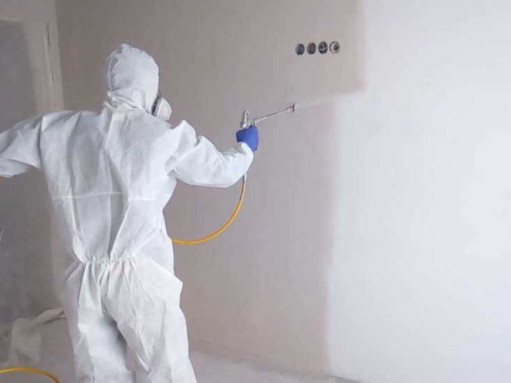 Pracownik w kombinezonie ochronnym i masce maluje ścianę natryskowo na biało; widoczne cztery otwory na gniazdka elektryczne nad świeżo pomalowaną powierzchnią.