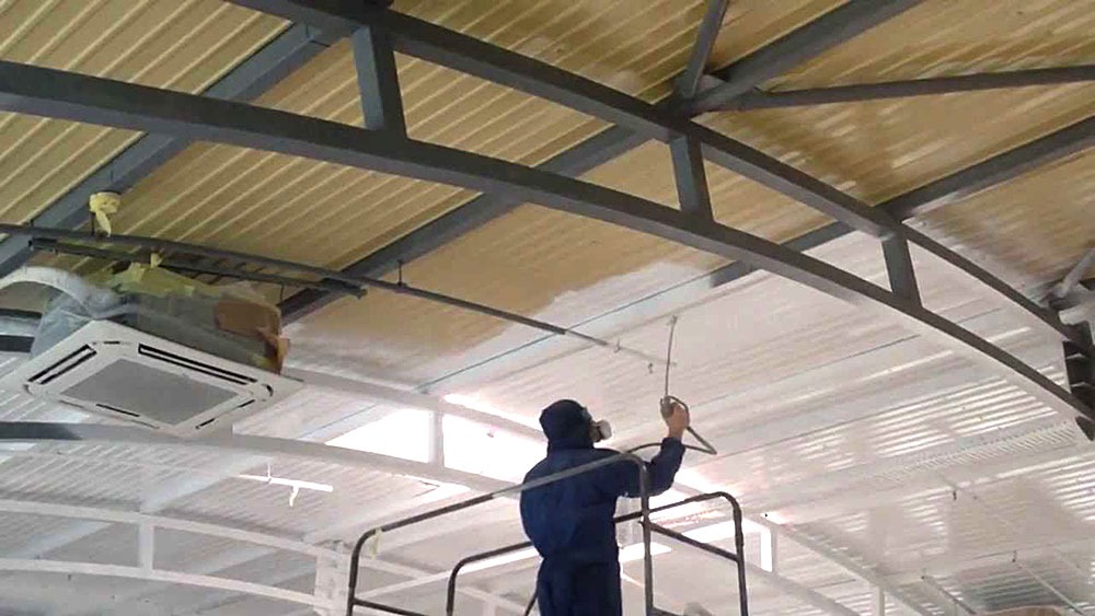 Pracownik w kombinezonie i masce ochronnej maluje natryskowo sufit w hali przemysłowej, widoczny niezabezpieczony klimatyzator.