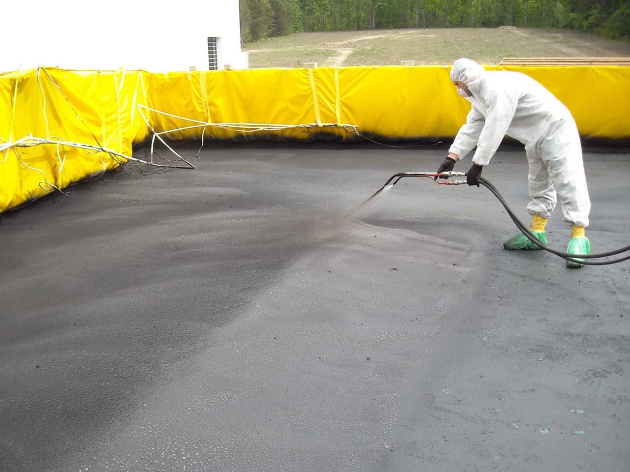 Pracownik w kombinezonie ochronnym natryskuje czarną substancję na płaską powierzchnię otoczoną żółtą barierą ochronną.