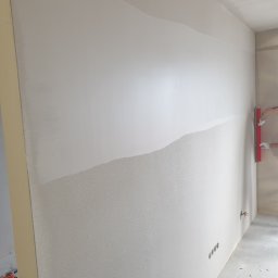 MPAKIET SP. Z O.O. - Ściana w trakcie wygładzania, widoczna część z nałożoną gładzią i pracownik z pacą w rękawicach, przygotowujący powierzchnię do malowania.