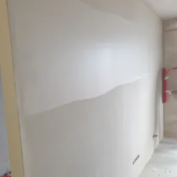 Ściana w trakcie wygładzania, widoczna część z nałożoną gładzią i pracownik z pacą w rękawicach, przygotowujący powierzchnię do malowania.