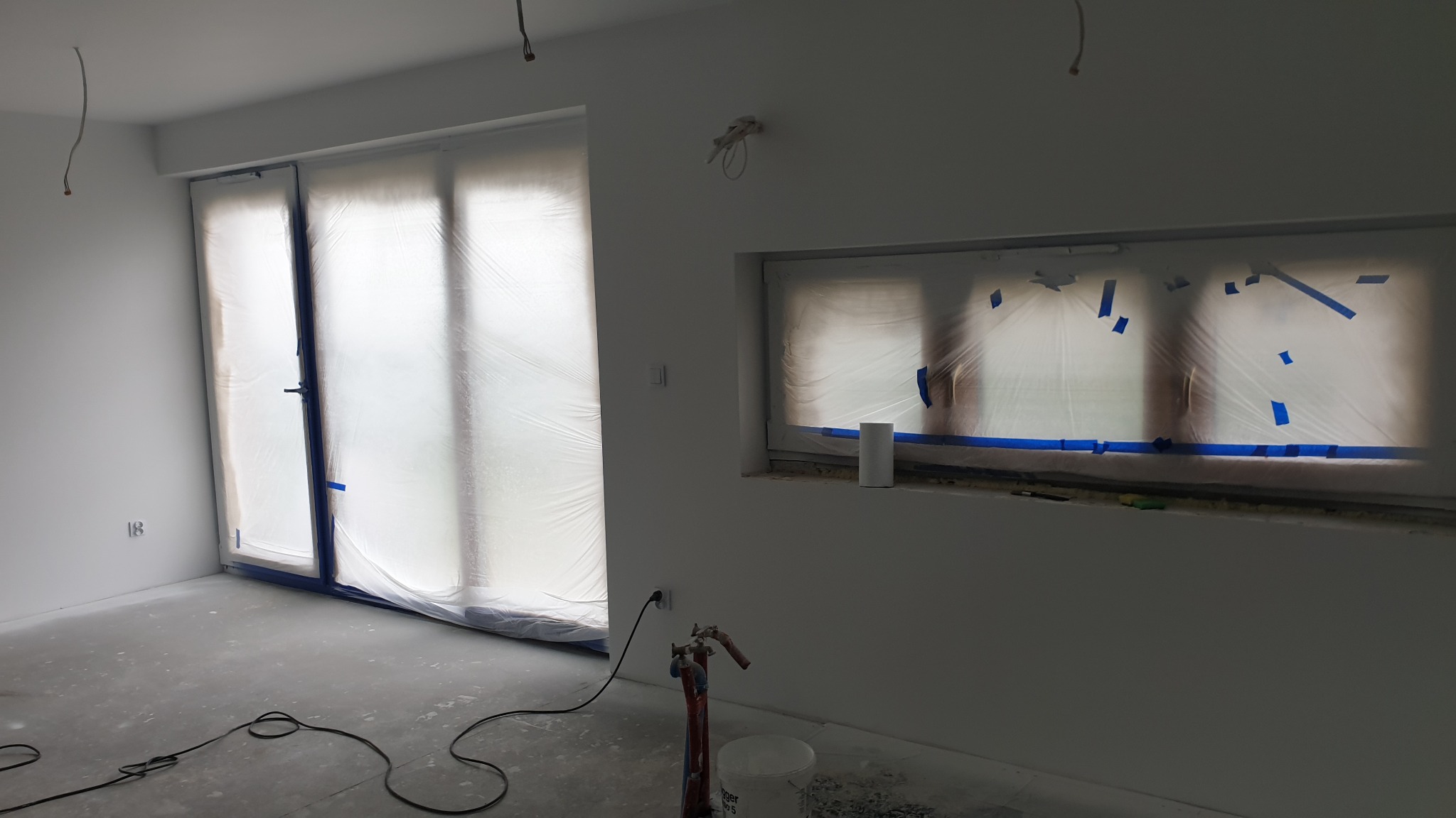 Przygotowanie do malowania: okna i drzwi zabezpieczone folią i taśmą malarską, ściany świeżo pomalowane na biało, widoczny agregat malarski i wiadro z farbą.