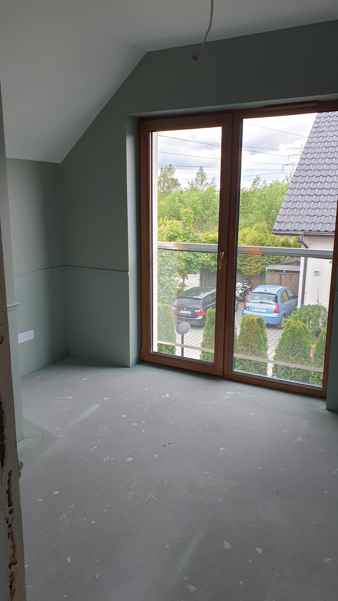 Pokój na poddaszu w trakcie prac przygotowawczych: świeżo pomalowane ściany w odcieniach szarości, surowa podłoga z widocznymi plamami, duże okno balkonowe z widokiem na podjazd z dwoma samochodami.