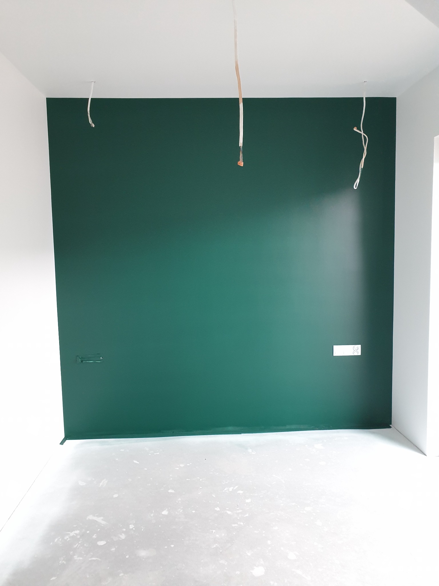 Świeżo pomalowana ściana na ciemnozielony kolor w pustym pokoju, widoczne wystające kable elektryczne i zakryte gniazdka, podłoga z resztkami farby.
