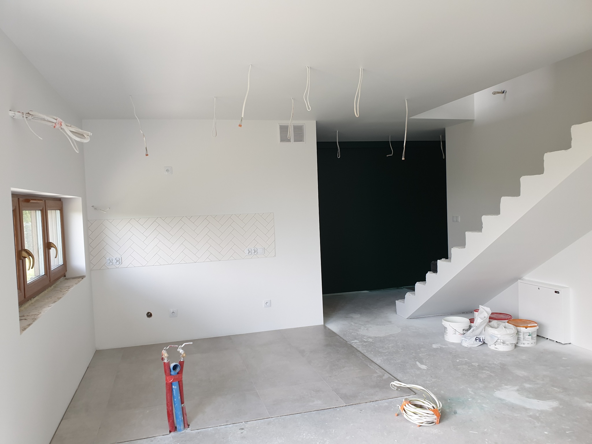 Wnętrze w trakcie prac wykończeniowych: białe ściany, częściowo ułożona szara podłoga z płytek, białe schody, widoczne puszki z masą szpachlową, wystające kable i rury instalacyjne.