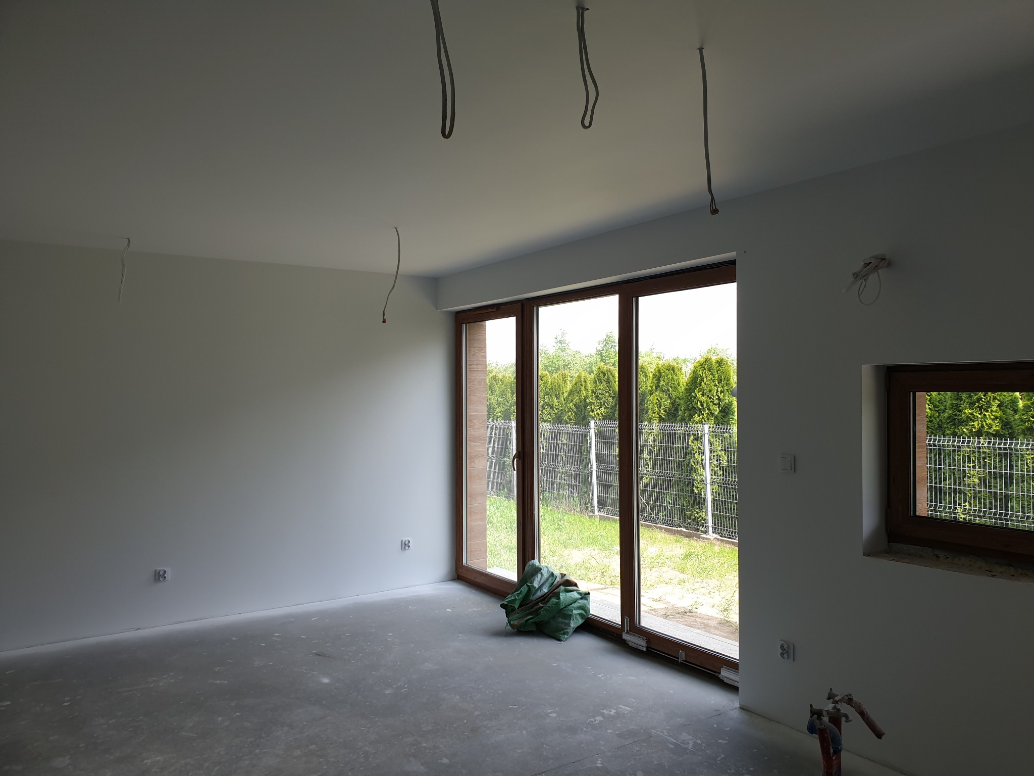 Pokój w stanie deweloperskim z dużym oknem balkonowym i małym oknem, widoczne wystające kable z sufitu i rury grzewcze przy ścianie, podłoga wylewka, ściany pomalowane na biało, na zewnątrz widok...