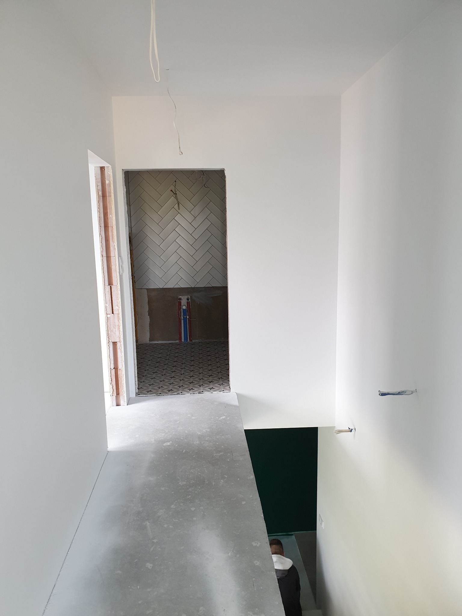 Korytarz w trakcie prac wykończeniowych: świeżo pomalowane białe ściany, surowa wylewka na podłodze, widoczne instalacje elektryczne wystające z sufitu, w tle łazienka z ułożoną białą glazurą...