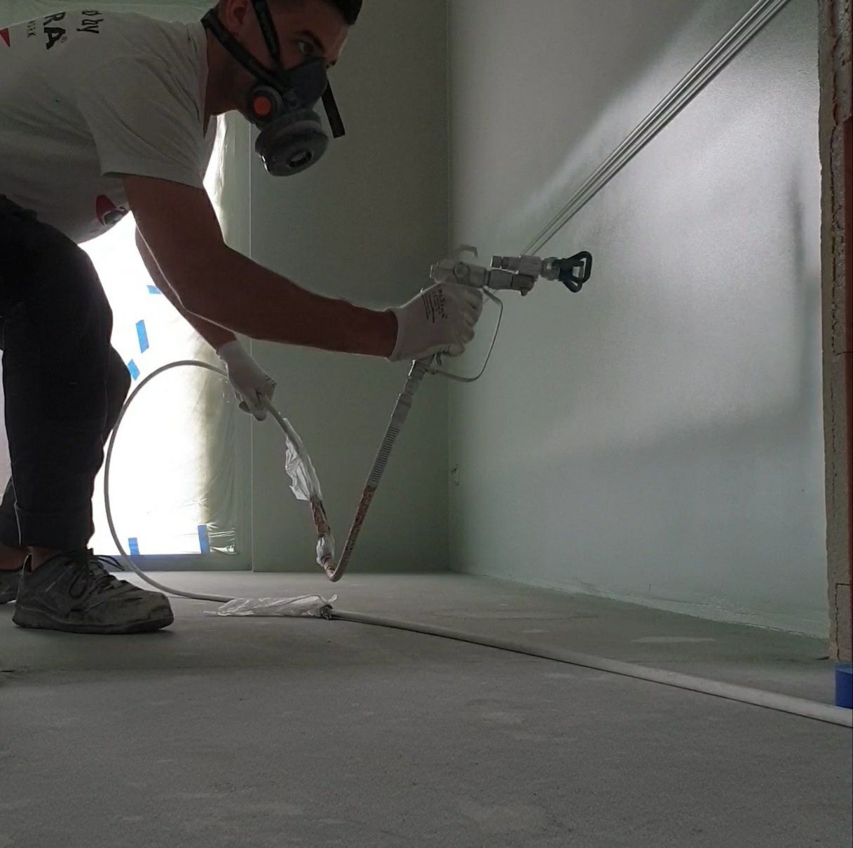 Pracownik w masce ochronnej maluje ścianę natryskowo, widoczny pistolet do malowania i przewód, zabezpieczona podłoga.