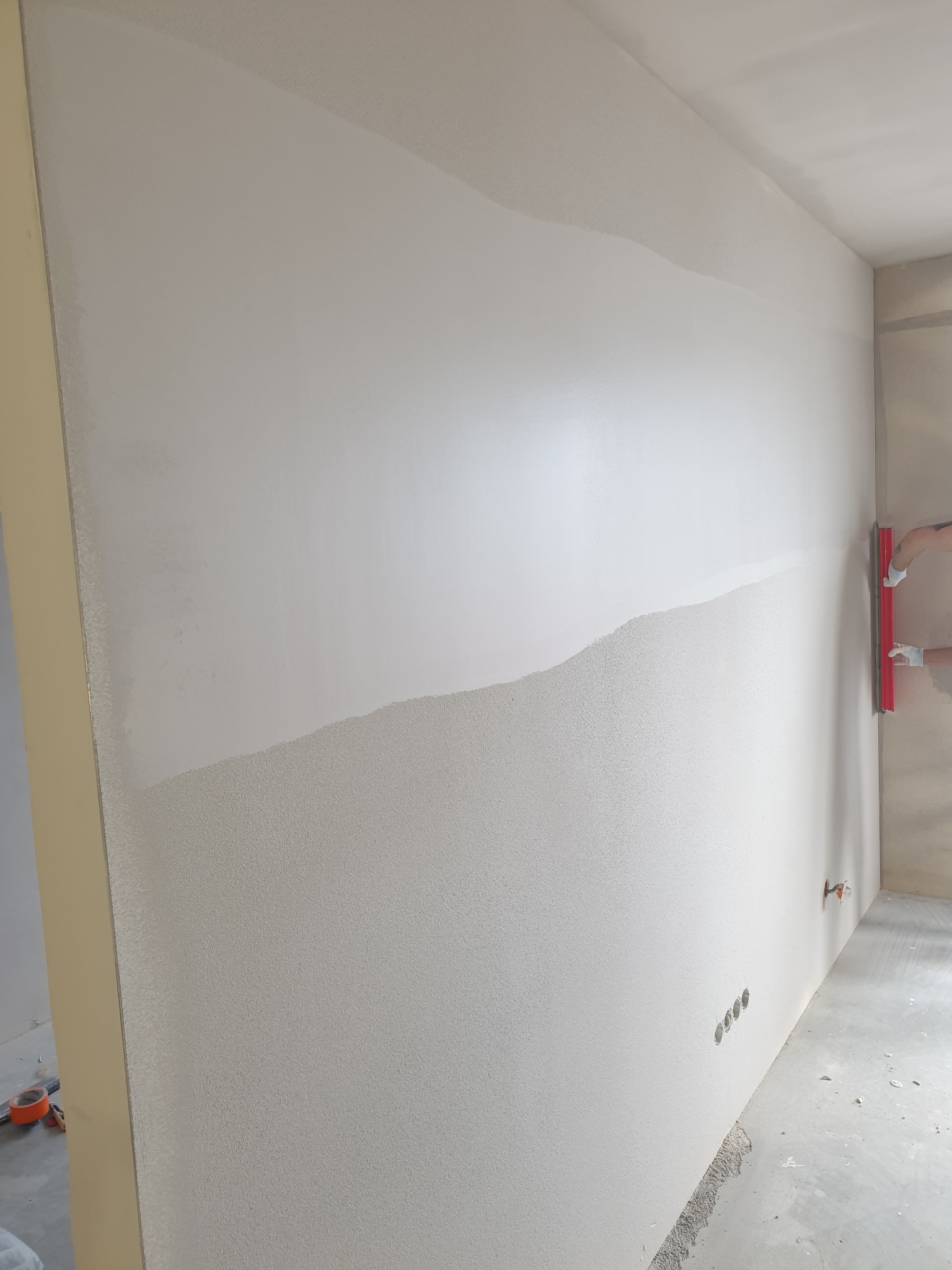 Ściana w trakcie wygładzania, widoczna część z nałożoną gładzią i pracownik z pacą w rękawicach, przygotowujący powierzchnię do malowania.