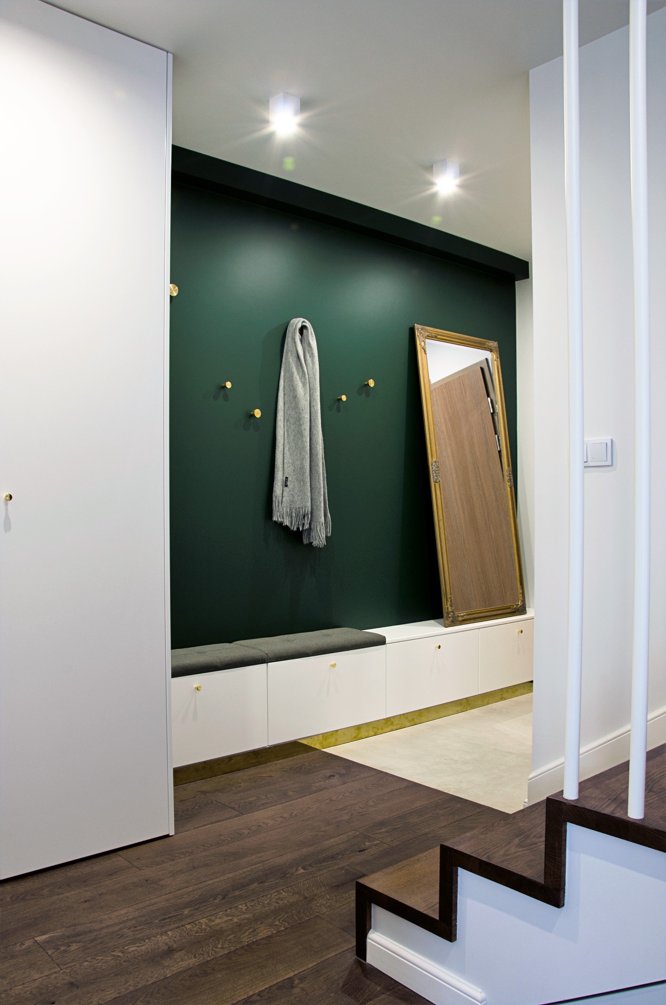 Nowoczesny przedpokój z ciemnozieloną ścianą, złotymi haczykami, szarym szalikiem, lustrem w złotej ramie, białą szafką z siedziskiem, drewnianą podłogą i schodami.