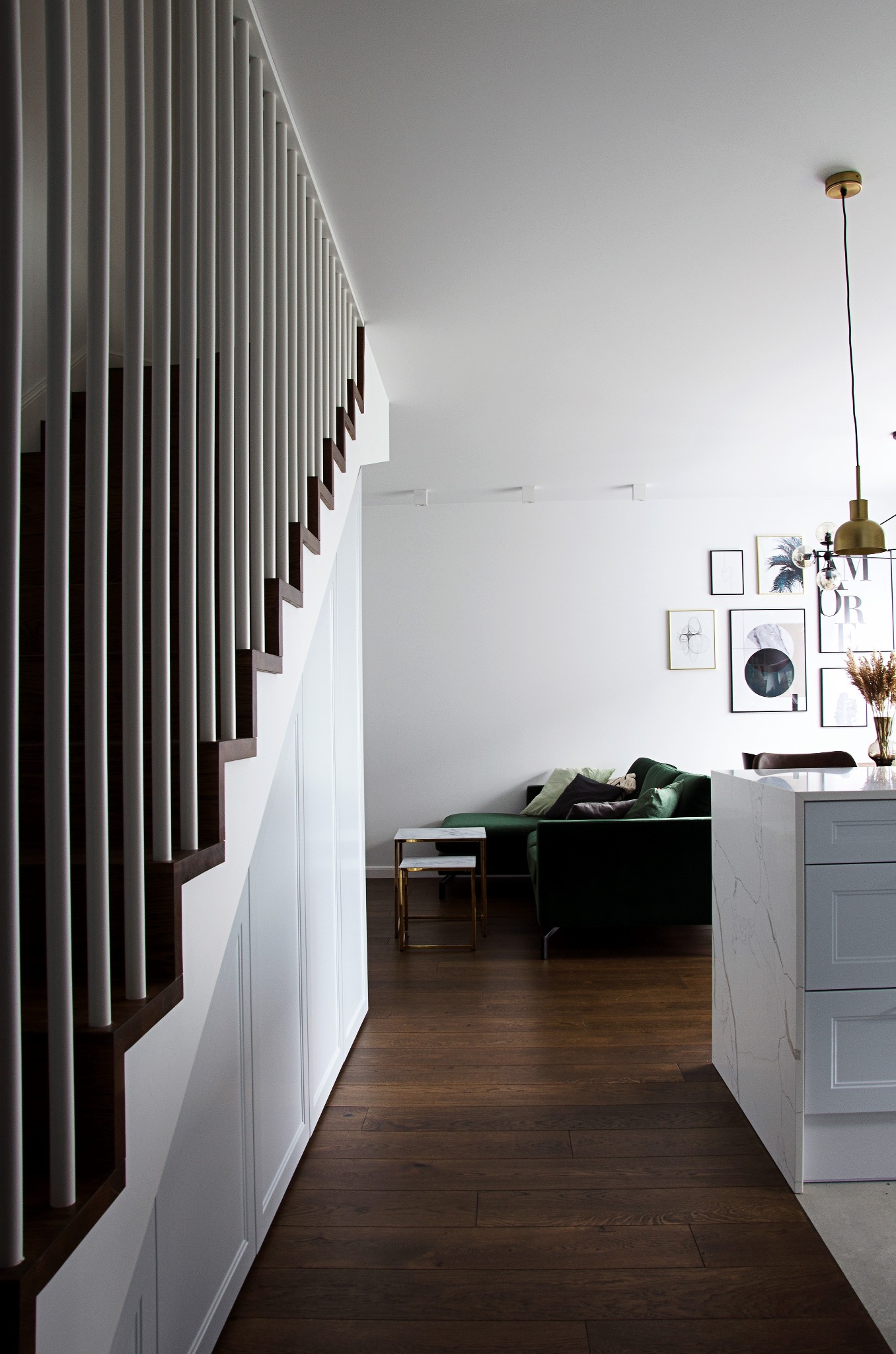 Nowoczesne wnętrze z drewnianą podłogą, schodami z białymi balustradami i zabudową pod schodami, w tle zielona sofa i galeria obrazów na ścianie, oświetlenie punktowe i wisząca lampa.