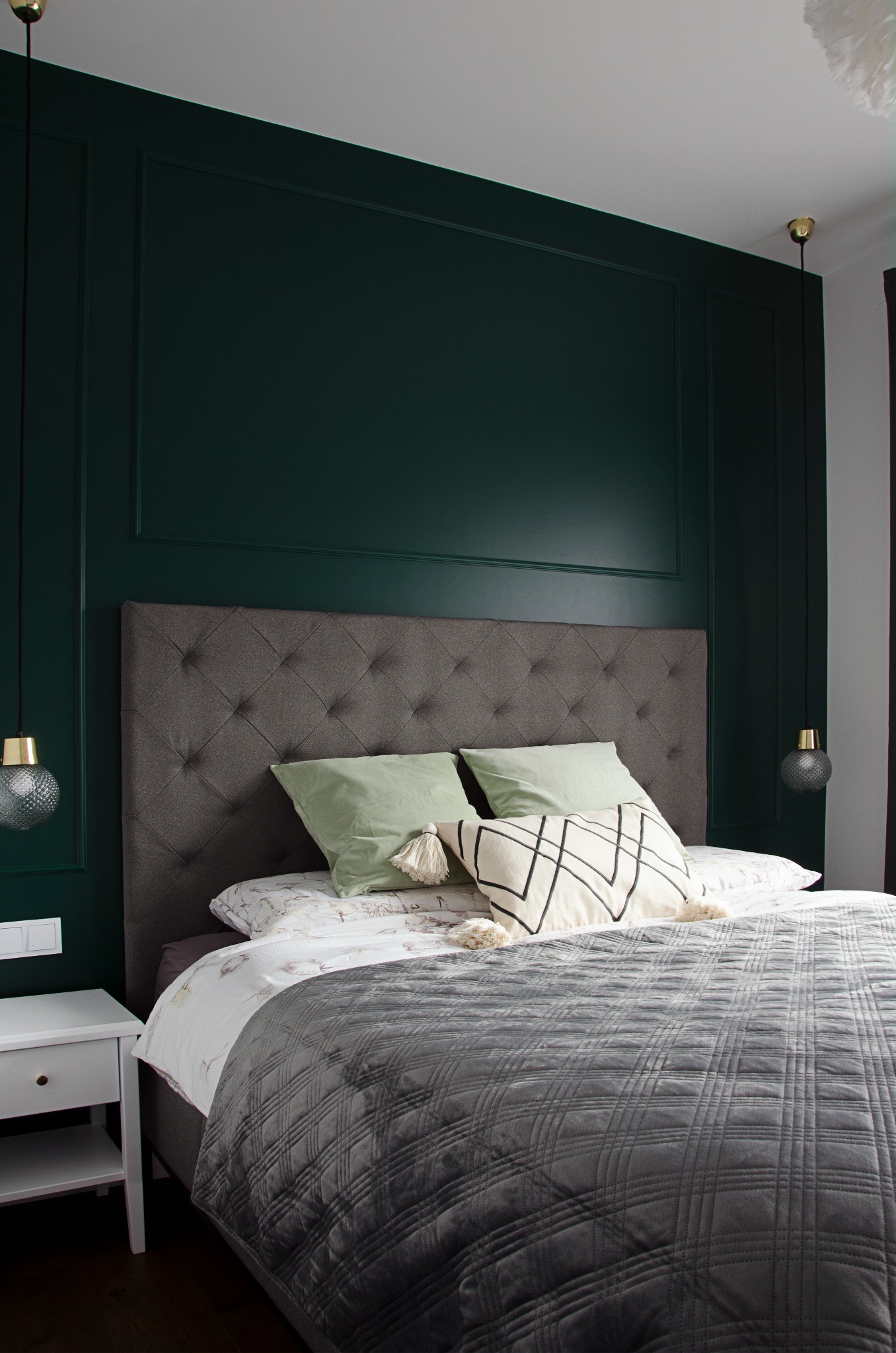 Nowoczesna sypialnia z ciemnozieloną ścianą w panelach, szarym pikowanym zagłówkiem i narzutą, lampkami nocnymi o kulistych kloszach i białą szafką nocną.