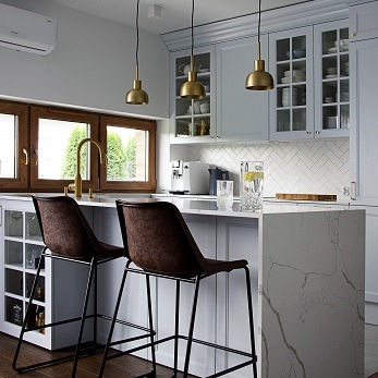Jasna kuchnia w stylu skandynawskim z drewnianymi oknami, białymi szafkami z przeszklonymi frontami, marmurowym blatem i wyspą. Nad blatem trzy złote lampy wiszące. Dwa brązowe krzesła barowe...