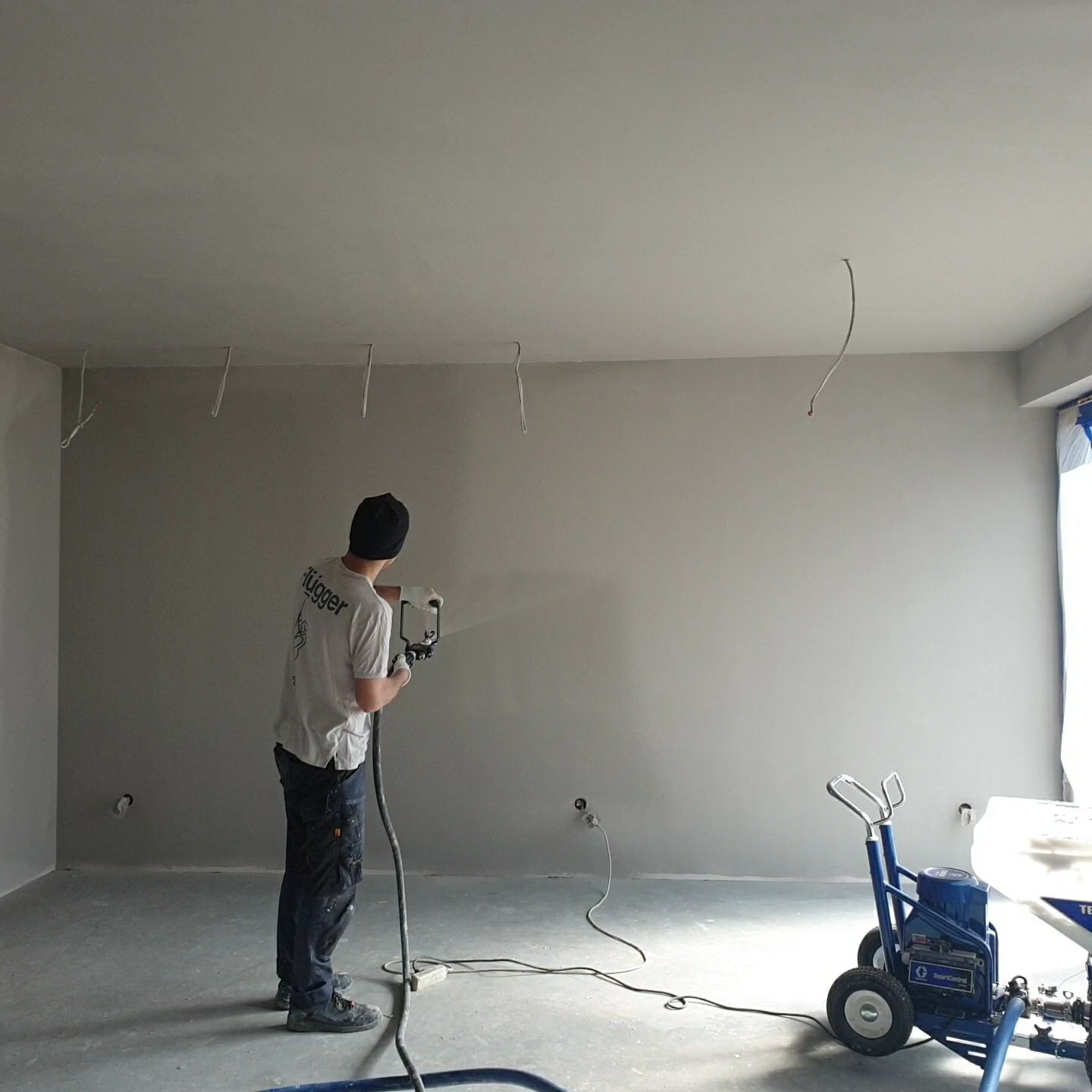 Pracownik w czapce maluje ścianę i sufit agregatem natryskowym, widoczne wystające przewody elektryczne, niebieski agregat malarski na kółkach.