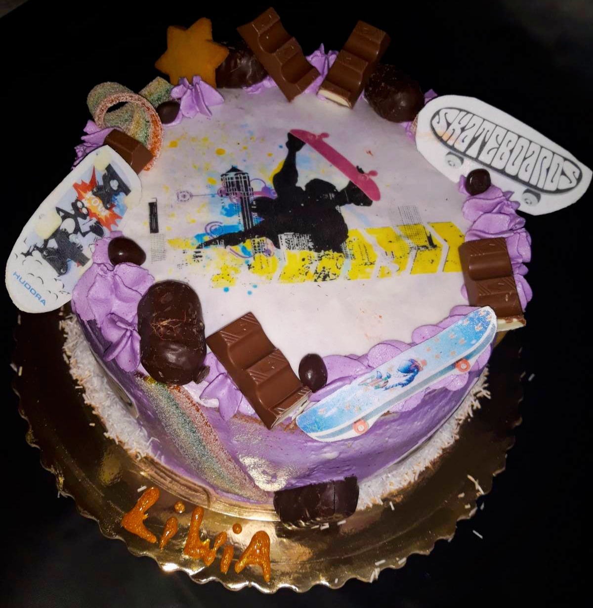 Tort urodzinowy z motywem skateboardingu, dekorowany fioletowym kremem, kawałkami czekolady, cukierkami i napisem 'Lilia' na złotej paterze.