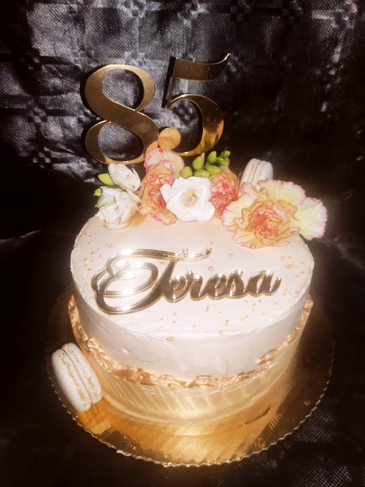 Dwupoziomowy tort urodzinowy z białym kremem, złotymi akcentami, kwiatami, makaronikami i topperem z liczbą 85 oraz napisem 'Teresa'.