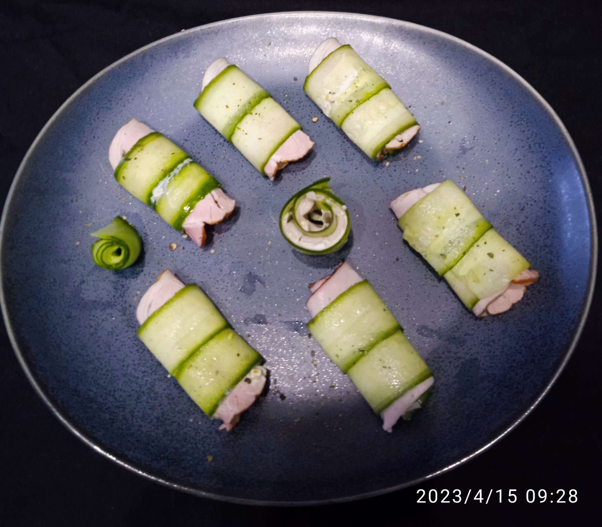 Dekoracyjne roladki z ogórka i szynki na ciemnym talerzu, jedna uformowana w rozetkę, posypane przyprawami.