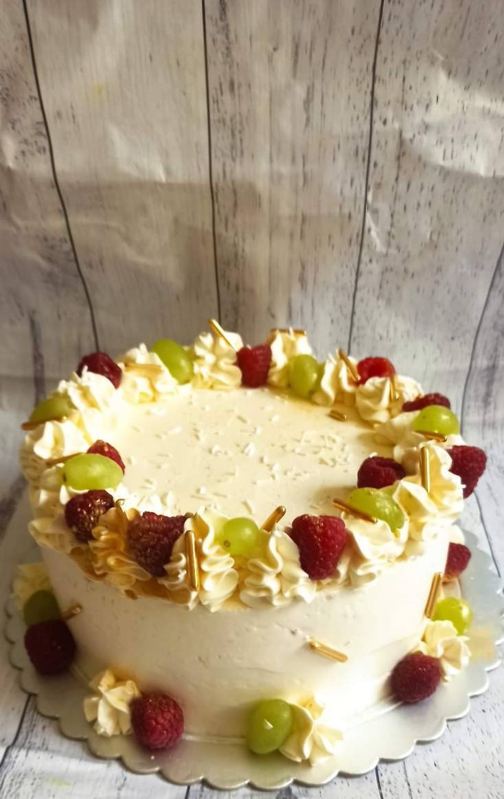 Tort śmietankowy udekorowany świeżymi malinami, zielonymi winogronami, kremowymi rozetkami i złotymi posypkami na srebrnej paterze, tło z desek.