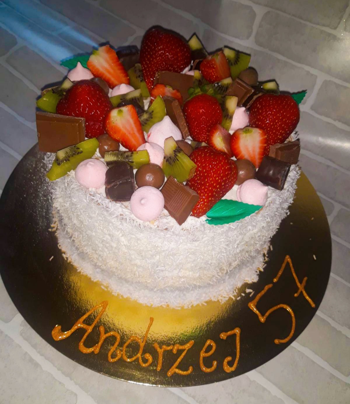 Tort urodzinowy z wiórkami kokosowymi, dekorowany świeżymi truskawkami, kiwi, kawałkami czekolady i małymi bezami, z napisem 'Andrzej 57' wykonanym złotym lukrem na złotej podkładce.