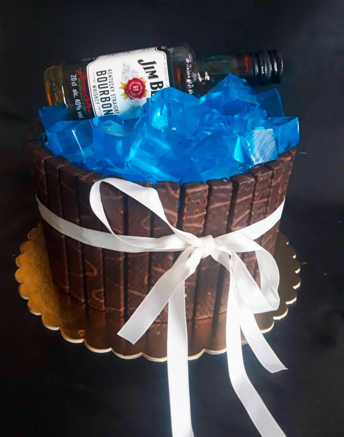 Tort obłożony batonikami, przewiązany białą wstążką, z niebieską galaretką w kostkach i butelkami alkoholu na wierzchu, na złotej podkładce.