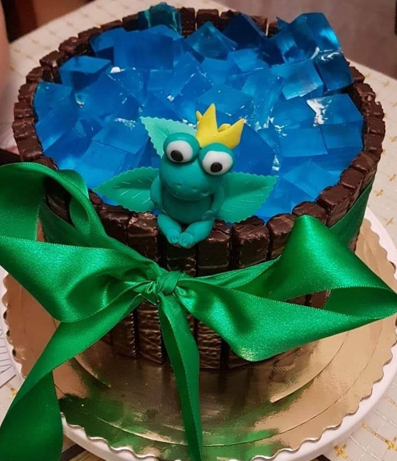 Okrągły tort zdobiony kostkami błękitnej galaretki, czekoladowymi batonikami, zieloną satynową wstążką i figurką zielonego żabiego króla.