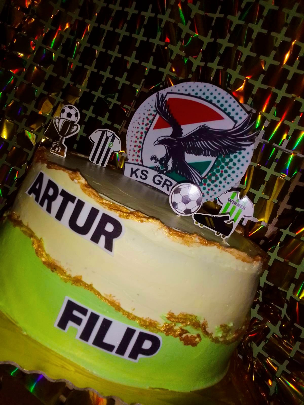 Tort urodzinowy w barwach klubu sportowego z imionami Artur i Filip, ozdobiony logo klubu, piłką nożną, pucharem i strojami piłkarskimi na tle błyszczącej folii.