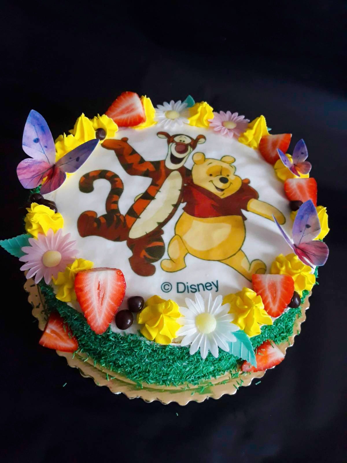 Tort z wizerunkiem Kubusia Puchatka i Tygryska, udekorowany truskawkami, motylami z opłatka i kremem w kolorach żółtym i zielonym, na czarnym tle.