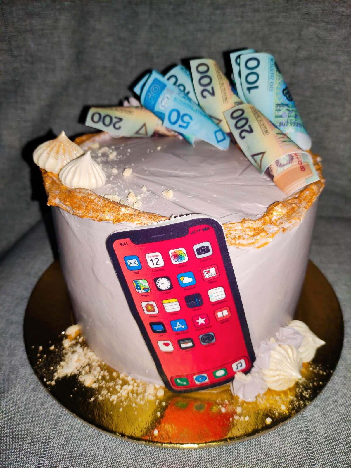 Tort urodzinowy z dekoracją imitującą banknoty i smartfon z ikonami aplikacji, na złotym podkładzie i szarym tle.