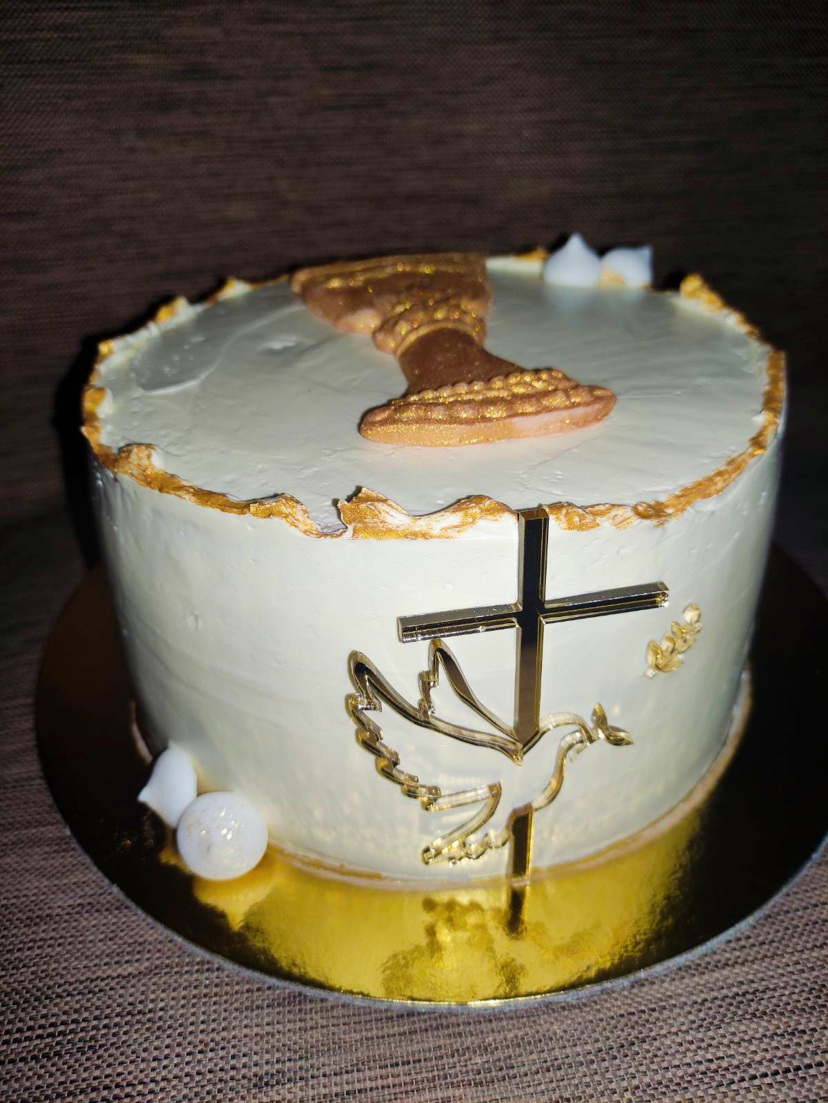 Tort komunijny z białym kremem, złotymi zdobieniami, hostią i krzyżem z gołębicą.