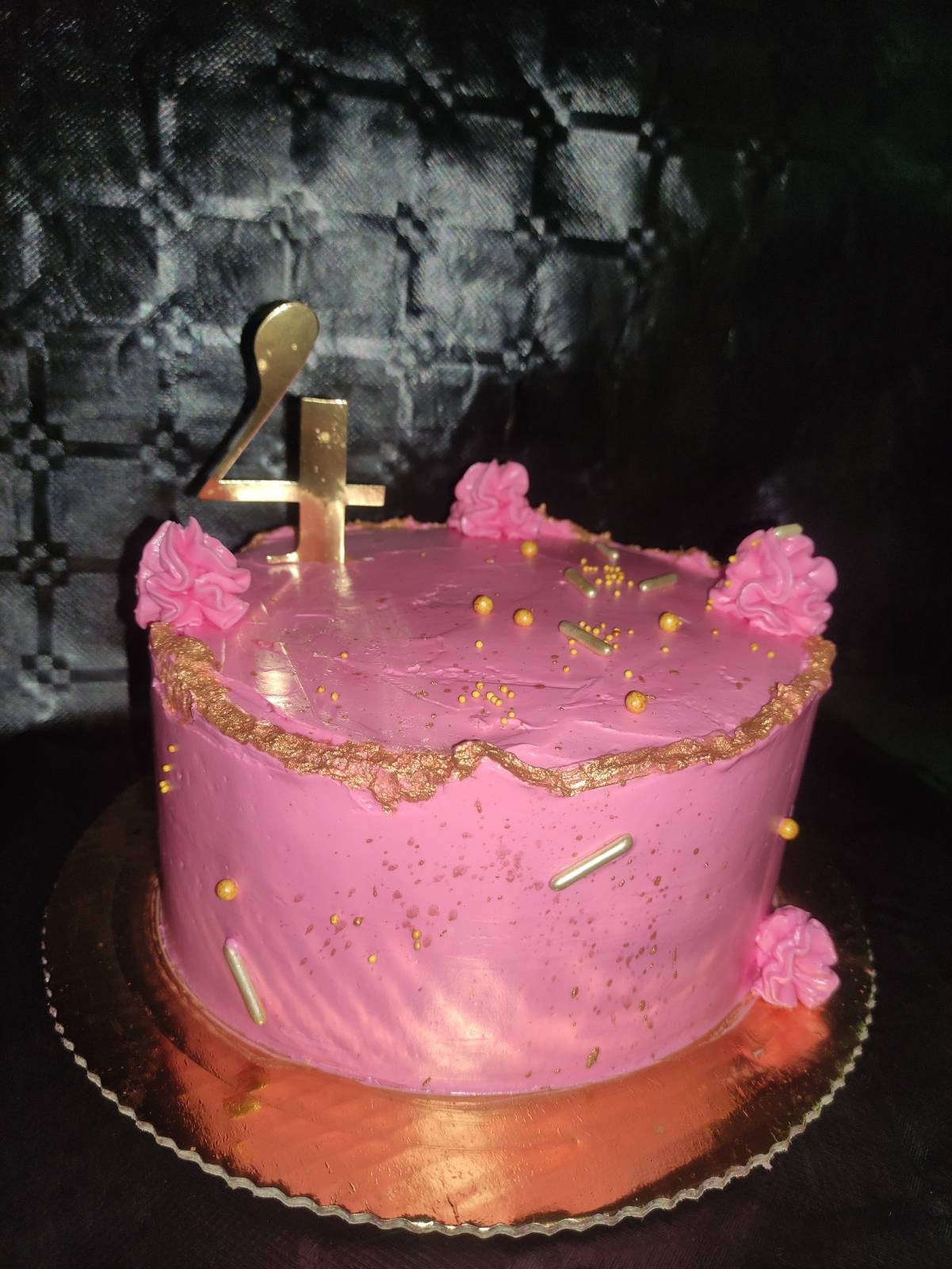 Różowy tort urodzinowy z dekoracjami z kremu i złotymi posypkami, na złotym podkładzie, z topperem w kształcie cyfry 4, na ciemnym tle.