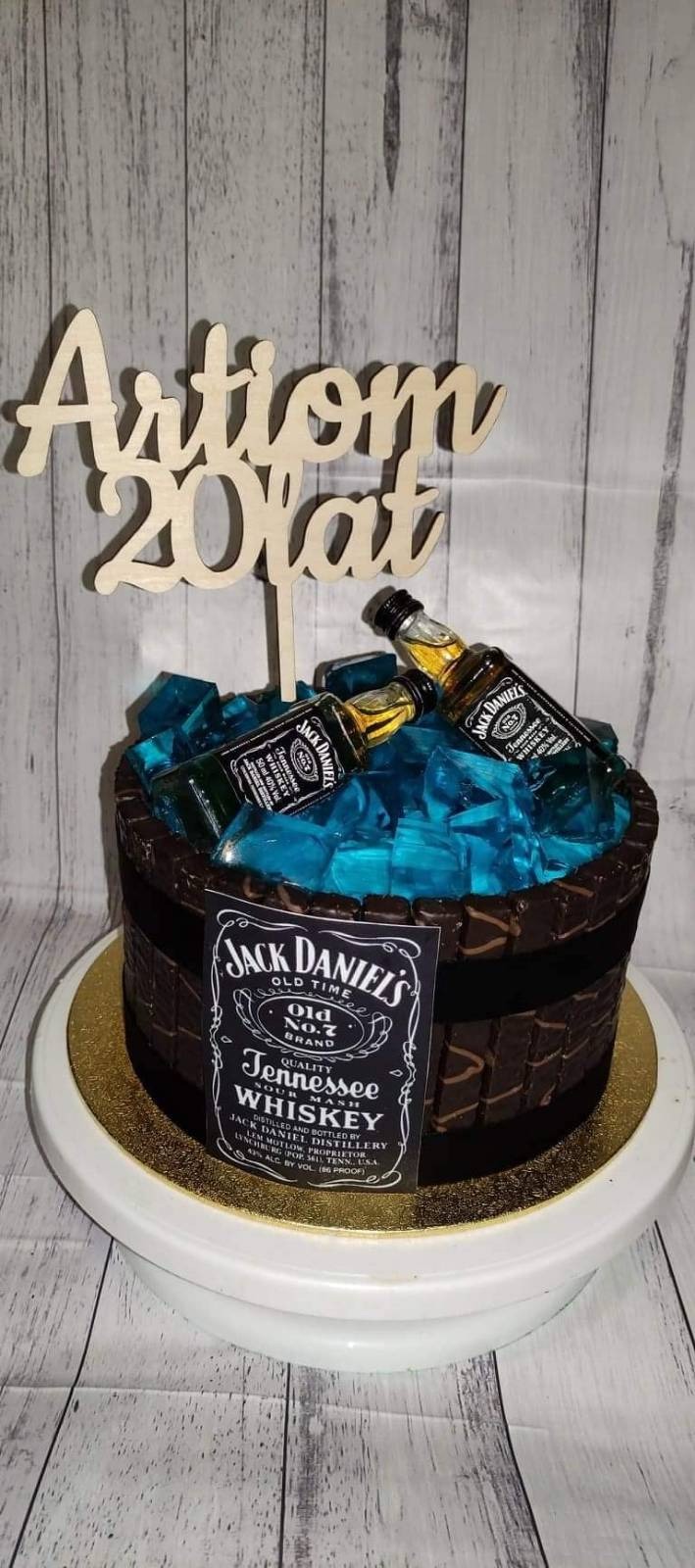 Tort w kształcie beczki z whiskey Jack Daniel's, wypełniony niebieskimi kostkami imitującymi lód, udekorowany miniaturowymi butelkami whiskey i drewnianym napisem 'Artiom 20 lat', umieszczony...