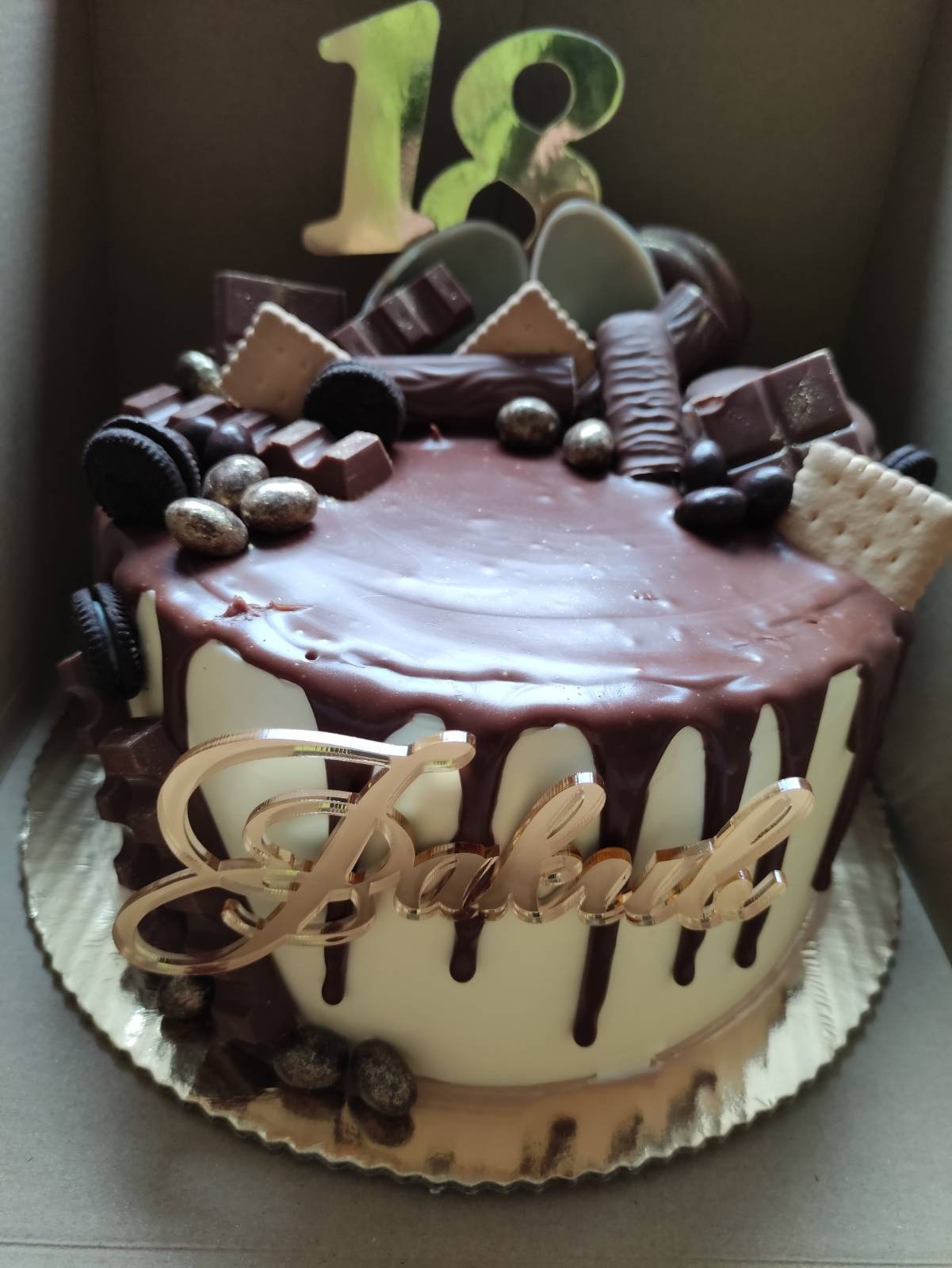 Tort urodzinowy z polewą czekoladową i dekoracjami z ciastek Oreo, batoników, złotych kulek i napisem 'Babcia' w złotym kolorze, zwieńczony topperem z liczbą 18 w złotym odcieniu.