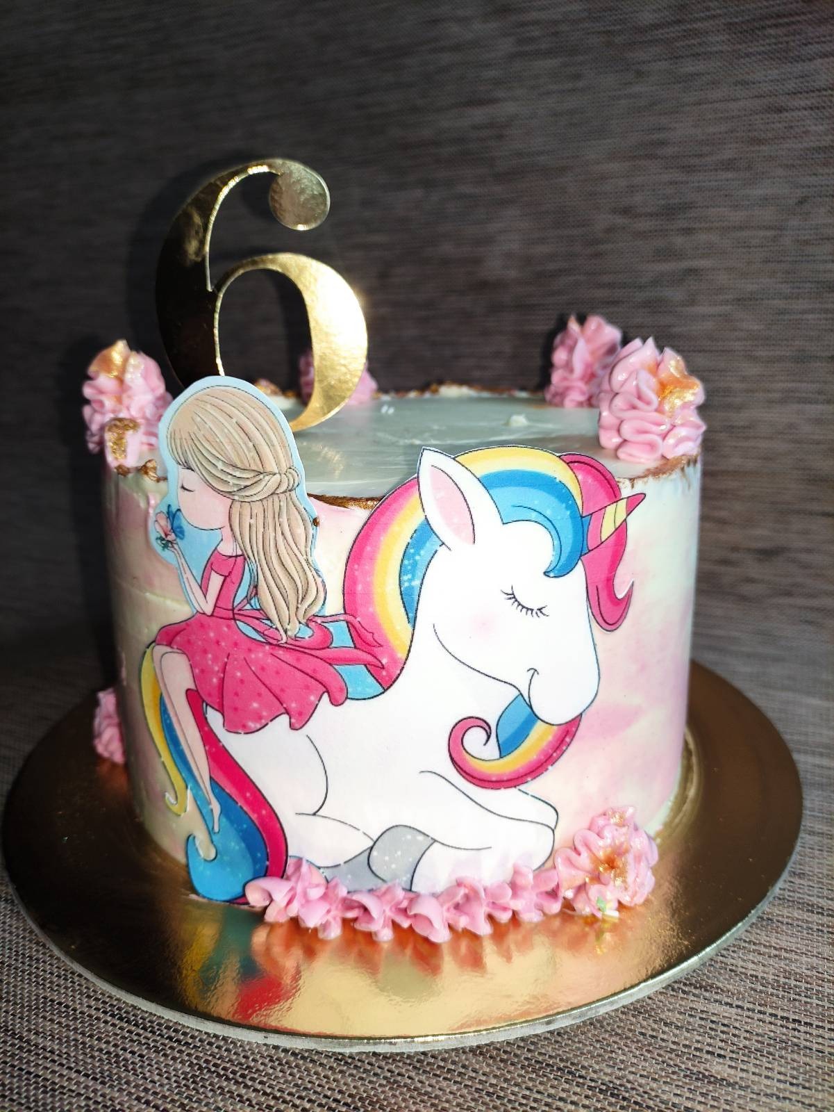 Tort urodzinowy dla dziecka z dekoracją przedstawiającą jednorożca i dziewczynkę, ozdobiony kremem w kolorze różowym oraz złotą cyfrą 6.