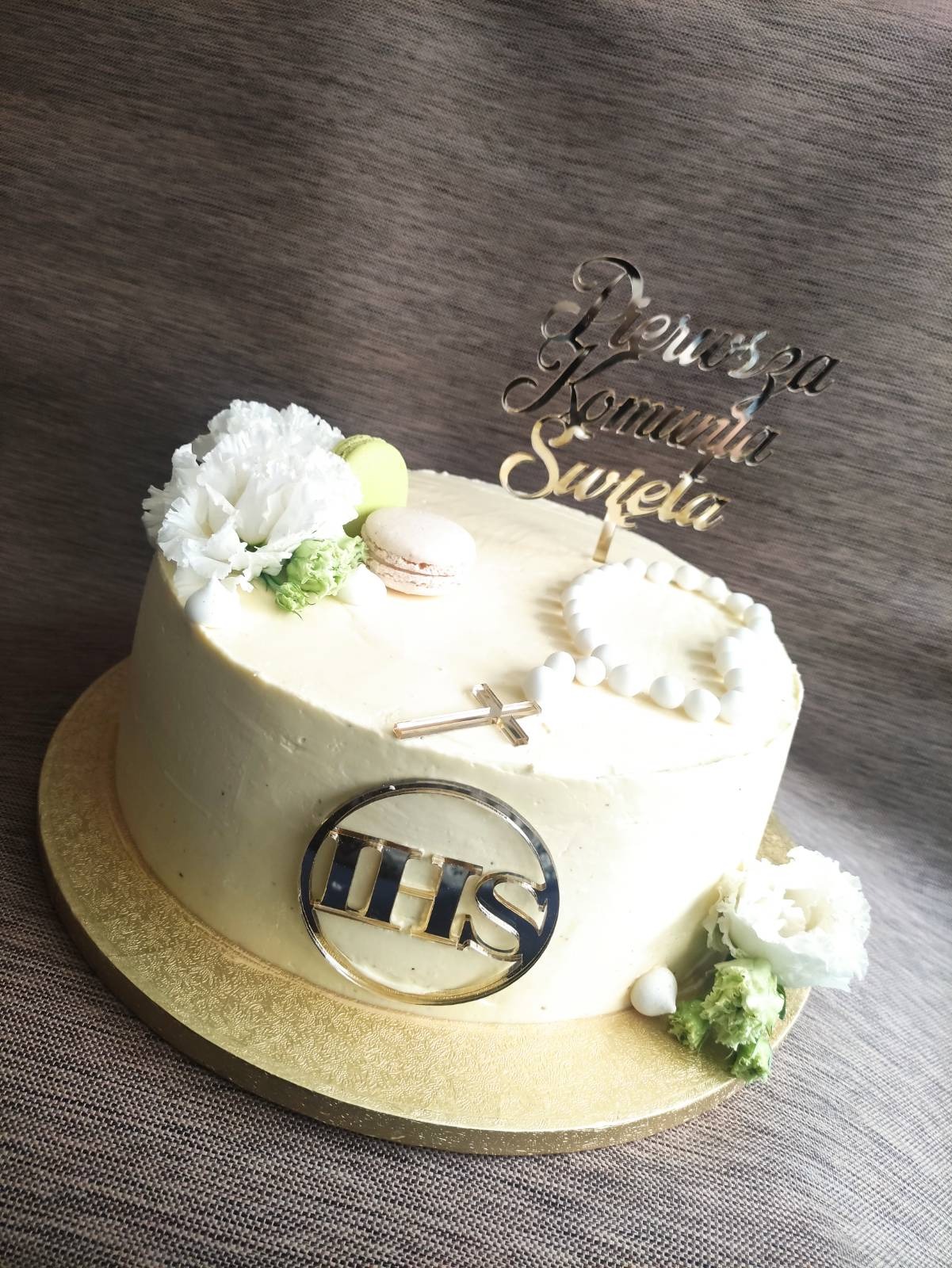 Tort komunijny z białym kremem, dekorowany kwiatami, makaronikami, różańcem z perełek, złotym krzyżykiem i napisem 'Pierwsza Komunia Święta' na złotym topperze oraz logiem IHS.