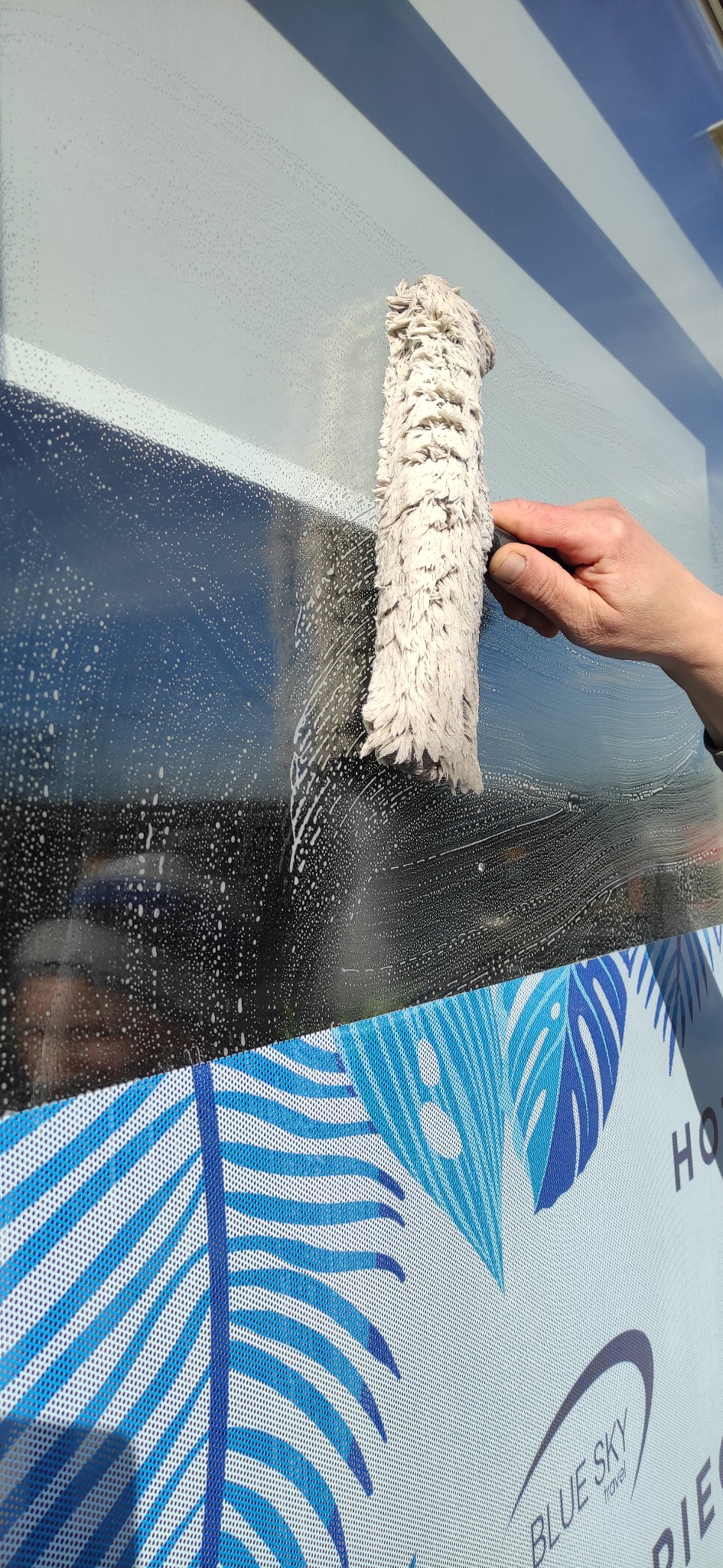 Ręka czyści okno z mydlin za pomocą mopa z frędzlami, widoczna reklama Blue Sky Travel z motywem liści palmowych w dolnej części szyby.