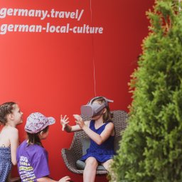 Martyna Kamzol - Dwie dziewczynki obserwują koleżankę w niebieskiej sukience, która siedzi w wiklinowym fotelu i używa zestawu VR na tle czerwonej ściany z napisem germany.travel/german-local-culture oraz zielonego...