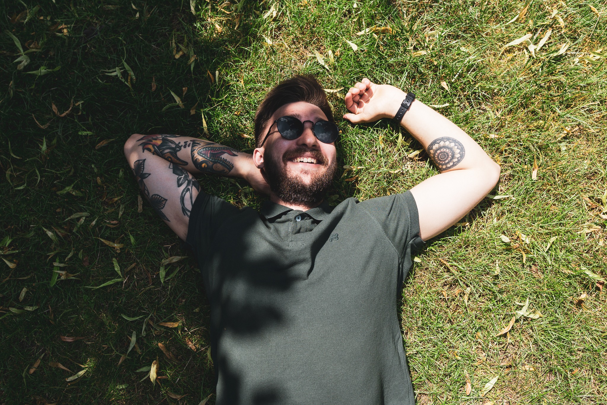 Mężczyzna z brodą i tatuażami leży na trawie w okularach przeciwsłonecznych, uśmiechając się w jasnym świetle słonecznym.
