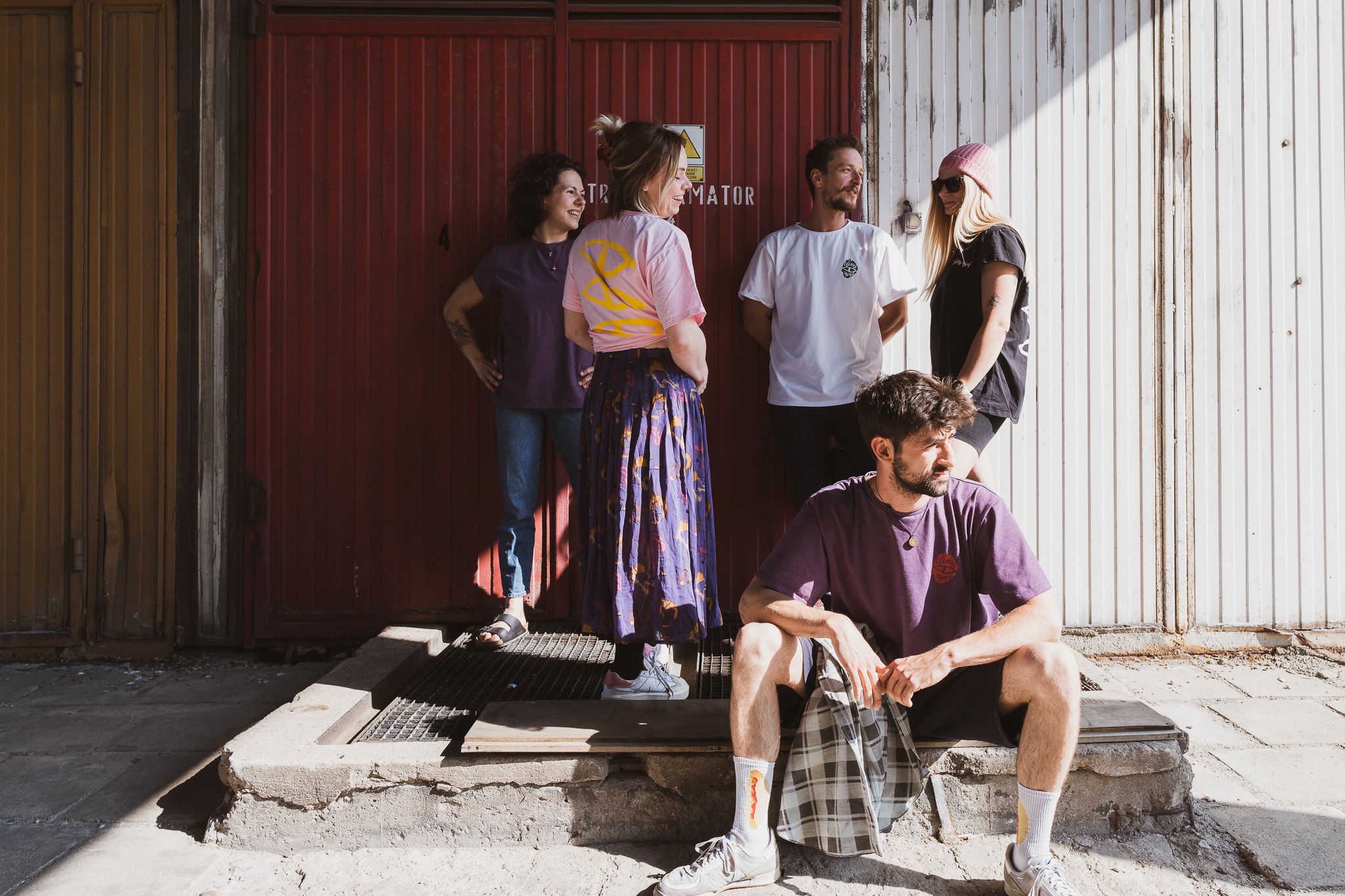Grupa pięciu młodych osób pozuje na tle kolorowych drzwi garażowych. Mężczyzna siedzi na betonowym murku, trzymając w rękach kraciastą torbę. Jedna osoba ma różową czapkę i okulary przeciwsłoneczne.